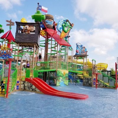 A colorful water playground featuring multiple slides, water cannons, and themed structures with characters, situated in a shallow pool area under a bright sky. A Texas flag is visible on top.