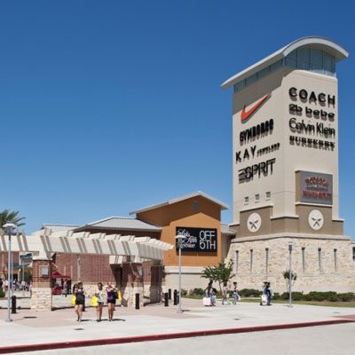 Shopping center with a tall tower displaying brand logos includes Coach, Nike, Calvin Klein, and more. Clear blue sky in the background with people walking near the entrance.