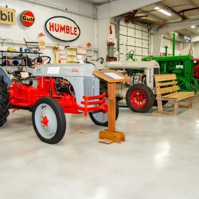 A display of vintage tractors in a museum setting, including a gray and red tractor in the foreground. Retro fuel company signs are mounted on the wall above.