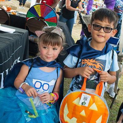 Two children in costumes hold goody bags at an outdoor event. One bag is shaped like a jack-o'-lantern. A table with a prize wheel is visible in the background.