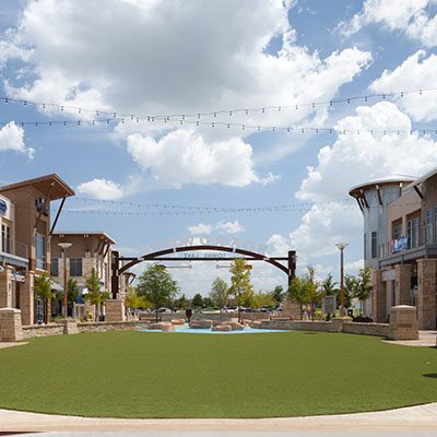 A green lawn and paved walkway sit between two rows of modern buildings under a partly cloudy sky, with decorative string lights overhead.