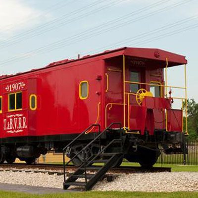A bright red caboose with yellow railings is parked on railroad tracks. The word "T.B.V.R.R." is painted on the side.