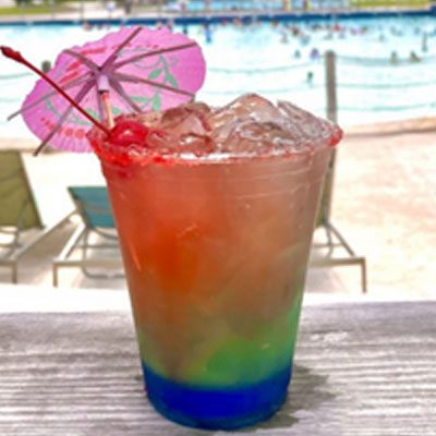 A colorful tropical drink in a clear plastic cup with a pink umbrella sits on a wooden surface. A swimming pool and lounge chairs are visible in the background.