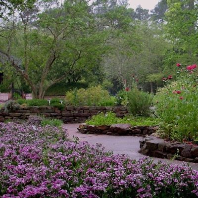 A garden path bordered by pink and purple flowers, with trees and greenery in the background, under an overcast sky.