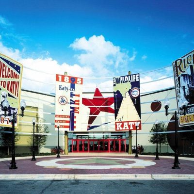 Front entrance of a building with large colorful banners displaying historical and local themes, such as "Welcome to Katy," "MKT," and "Rice." The sky is clear with a few clouds, and there are trees nearby.