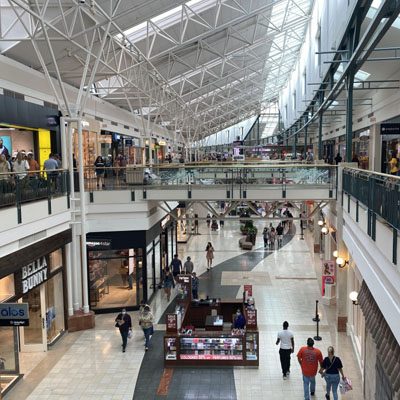 A view of a multi-level shopping mall with people walking, shops on both sides, and a central kiosk. The mall has a bright, spacious design with a high, glass-paneled ceiling.
