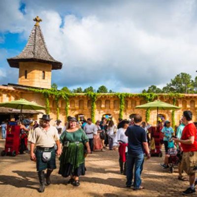 A crowd of people in diverse attire walk through an outdoor festival with a stone building and tower in the background.