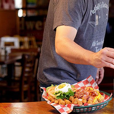 Person reaching for a basket of food containing a burger with toppings and fried items on a checkered paper, in a restaurant setting.