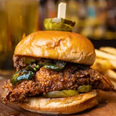 A spicy chicken sandwich with jalapeños and pickles on a brioche bun, skewered with a toothpick. Fries are visible in the background.