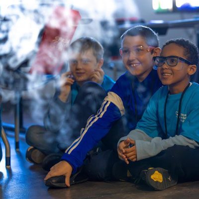 Three children sitting on the floor, attentively watching something, with smoke or mist partially obscuring their view. They appear engaged and are wearing blue shirts.