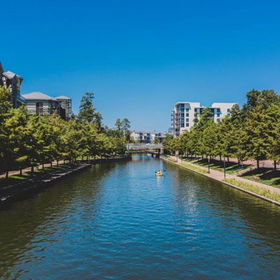 A peaceful canal lined with trees and buildings under a clear blue sky, with a person kayaking in the center.